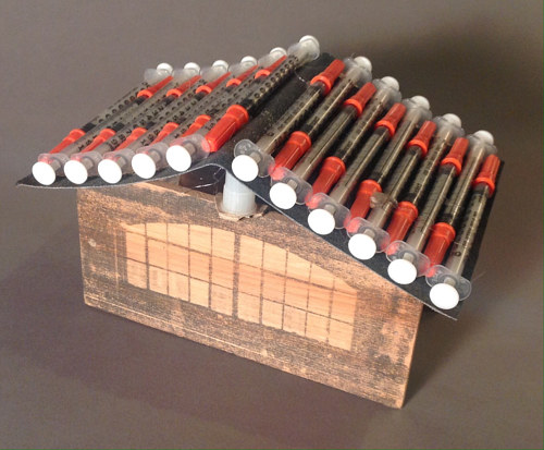 A sculpture of a small house with syringes making up the roof